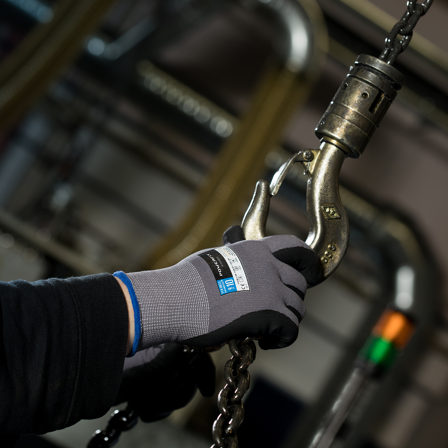 En handskbeklädd hand förbinder en metallkrok till en kraftig kedja i industriell miljö med Montagehandskar Powerfit® 12 st Granberg.