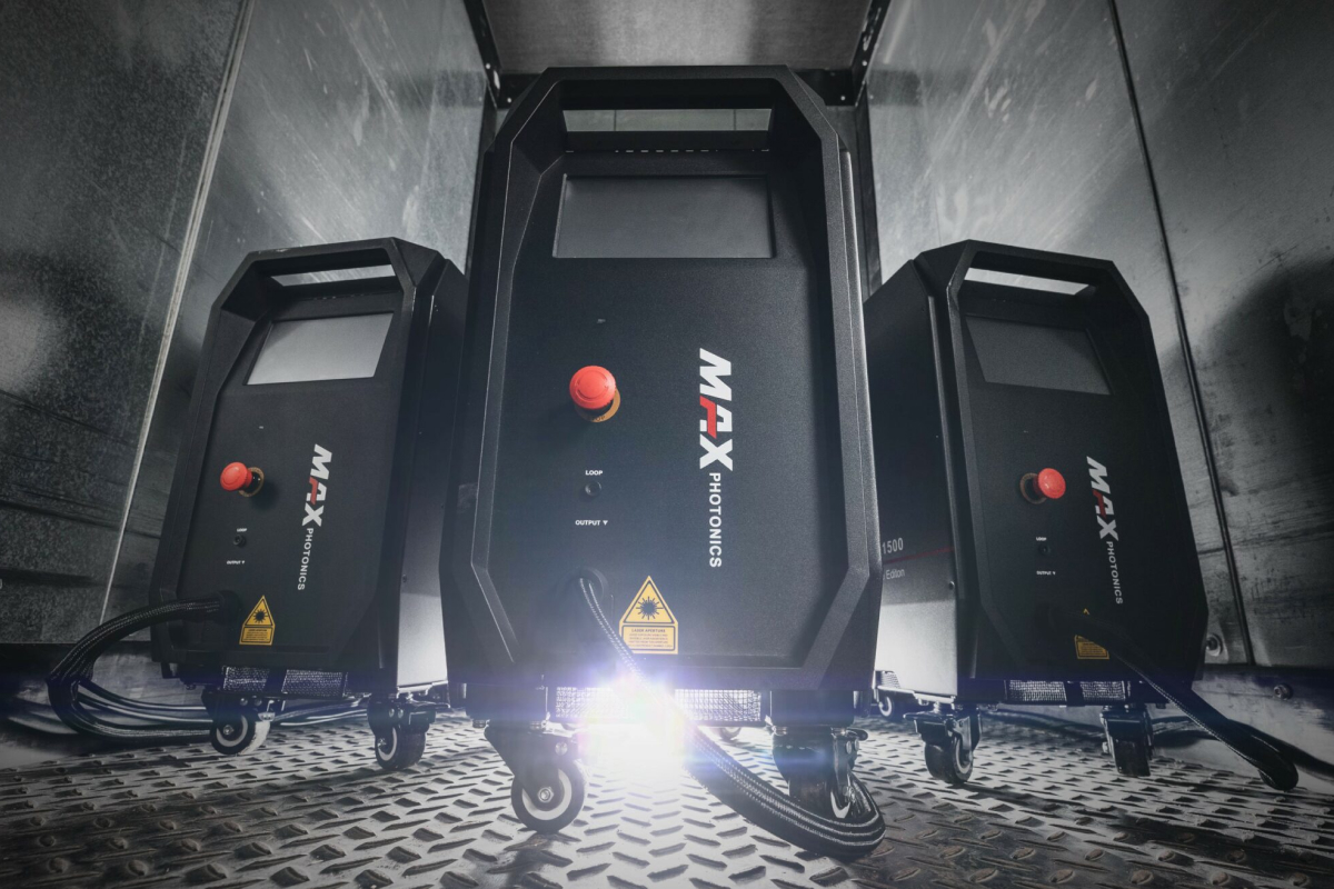 Tre MAX PHOTONICS industrimaskiner med kontrollpaneler och kablar på ett gallergolv i en svagt upplyst miljö.